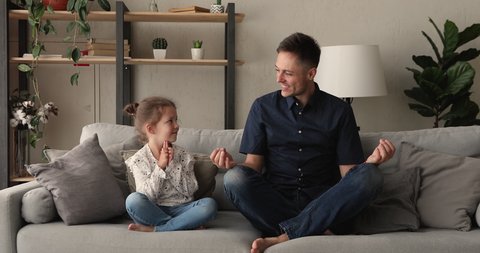 Tranquil father and little 5s daughter seated cross-legged on comfy couch closing eyes do together meditation asana, spiritual powerful practice. Teach kid manage emotions, healthy lifestyle concept