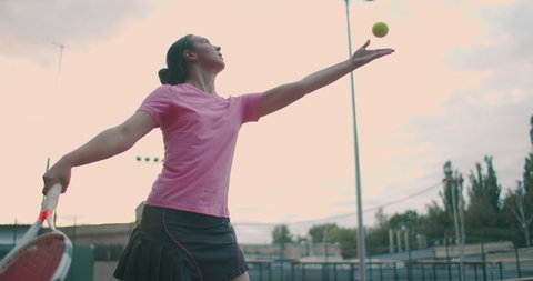 Slow motion: female sportsman during her practice. A close-up of a girl athlete serves the tennis ball. Young woman is hitting the ball with her tennis racket at sunset