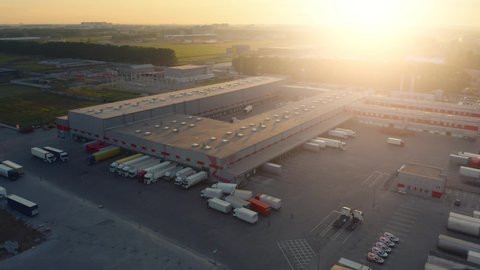 Aerial view of the large logistics park - loading hub with a lot of semi-trailers trucks standing at the ramps of the warehouse to load / unload goods at sunset.