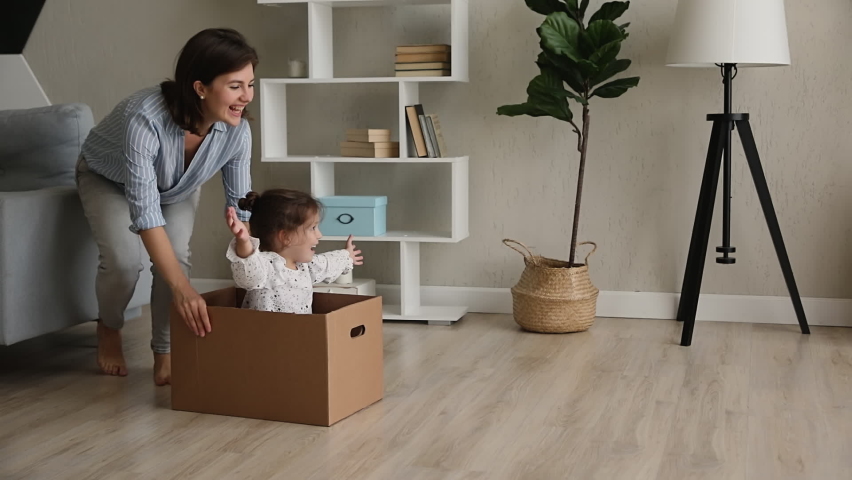 Young happy mother riding cute preschool daughter while she sit inside of big cardboard box, family laughing play active game at modern warm house celebrate relocation day to new comfort home concept Royalty-Free Stock Footage #1062680161