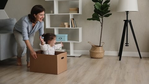 Young happy mother riding cute preschool daughter while she sit inside of big cardboard box, family laughing play active game at modern warm house celebrate relocation day to new comfort home concept