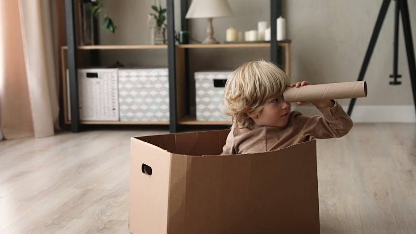 Cute little kid boy sit inside of cardboard box holding paper tube looking into distance imagines himself discoverer. Preschool curious child holding carton handmade binoculars play game alone at home Royalty-Free Stock Footage #1062681715