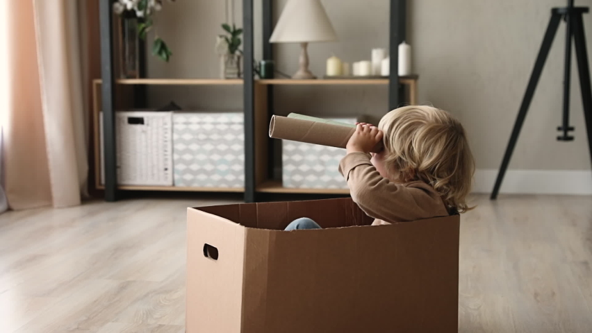Cute little kid boy sit inside of cardboard box holding paper tube looking into distance imagines himself discoverer. Preschool curious child holding carton handmade binoculars play game alone at home Royalty-Free Stock Footage #1062681715