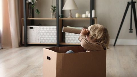 Cute little kid boy sit inside of cardboard box holding paper tube looking into distance imagines himself discoverer. Preschool curious child holding carton handmade binoculars play game alone at home