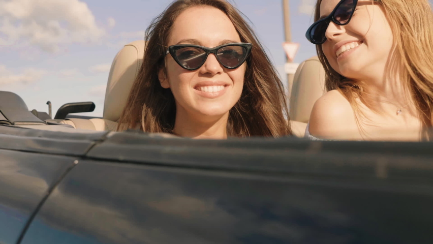 Portrait of two young beautiful and smiling hipster girls in convertible car. Sexy carefree women driving cabriolet. Positive models riding and having fun and raising hands