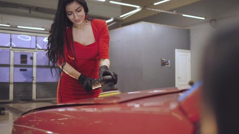 Portrait of a sexy auto center worker. The concept of an attractive girl from the detailing center. The girl in red polishes the car. Restoration of color and gloss of paint.