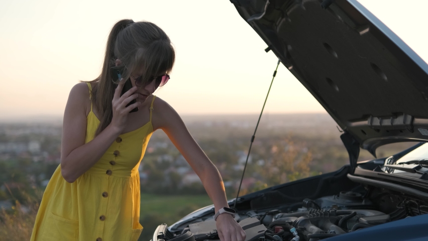 Stranded female driver talking on cell phone standing near her vehicle with open hood having engine problems.
