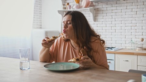 Junk food, eating disorder. Overweight woman with big appetite eating pizza, sitting at kitchen at home.
