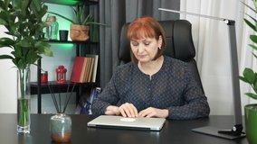 Motivated middle-aged businesswoman is opening laptop computer and starting to work at her office work desk. Female office worker is sitting in office in front of notebook, preparing to work