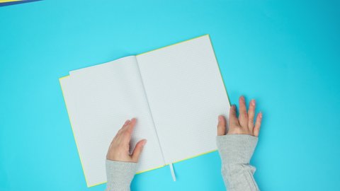 two female hands turn over the pages of a yellow notebook with blank white sheets on a blue background, top view
