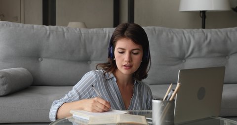 Young woman sit in living room at coffee table listens teacher through wireless headphones writing notes in copybook learn english remotely using laptop video call app. Modern tech, e-learning concept