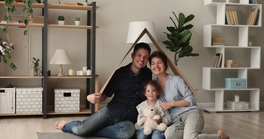 Happy beautiful couple with daughter sit on floor in domestic room under carton roof smile look at camera, take care and protection of kid together, new home, ownership, insurance, bank loan concept Royalty-Free Stock Footage #1062694678