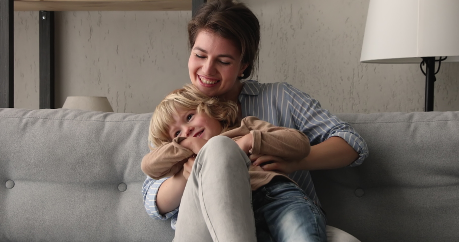 Little cheerful son sit on mum laps enjoy funny active playtime, laughing mom babysitter play tickle small boy sit on sofa in living room. Happy motherhood, leisure activity with child at home concept Royalty-Free Stock Footage #1062694714