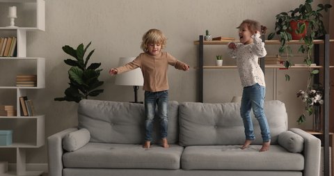 Small active boy and girl jumping barefoot on sofa in cozy warm living room, charming preschool children siblings or friends having playful funny mood play together at home, kids hyperactivity concept