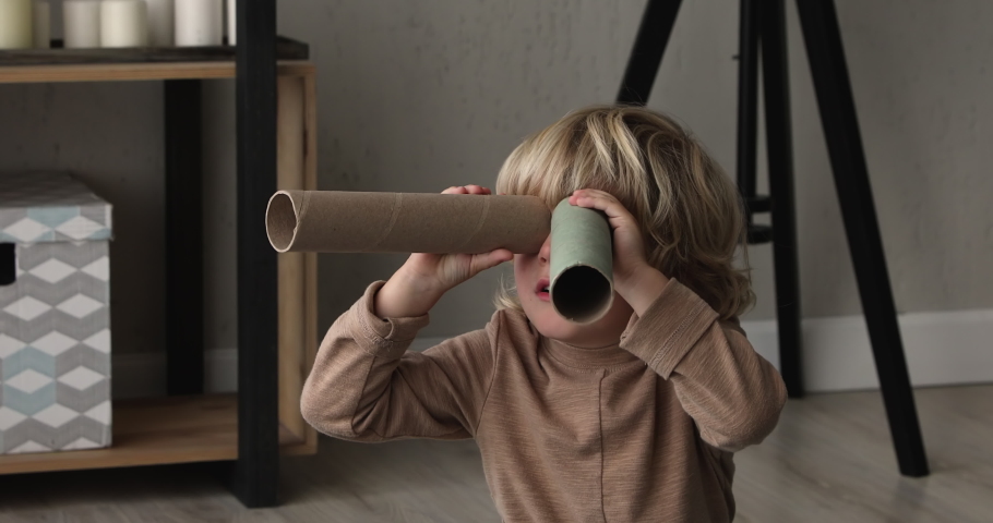 Close up of small cute boy sit in living room play alone holding cardboard tube like binocular watching into distance, preschool kid imagines look in telescope. Dreams, aspiration, imagination concept