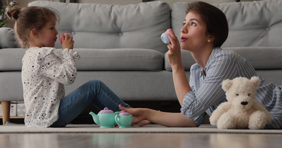 Loving mom playing with 5s daughter in warm living room, hold teapot pours coffee into tiny toy mug imagining like drink tea enjoy girlish talk at home. Playtime, Happy Mother Day celebration concept | Shutterstock HD Video #1062695917