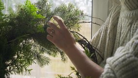Young woman in white sweater puts golden toy ball on wreath for Christmas holiday with green fir twigs standing near large window with curtains closeup - Powered by Shutterstock - Get 15% off with code: PIKWIZARD15