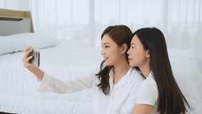 Group of young beautiful Asian woman in white casual outfit using front camera from smartphone to take a selfie with happy and joyful emotion in modern bedroom