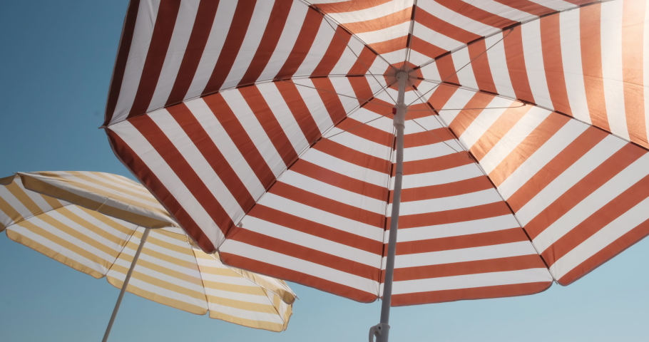 Two side by side striped umbrellas on a beach on a sunny perfect chilling day. Covering the sun, creating a nice shadow for relaxation and peacefulness in front of a sea ocean water and blue ideal sky Royalty-Free Stock Footage #1062703705