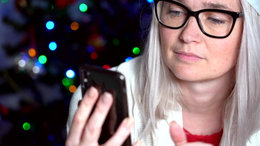 Close-up view  of happy woman in red santa hat sending air kiss over christmas tree lights background. Woman looking camera and waving greeting hands. 4k video