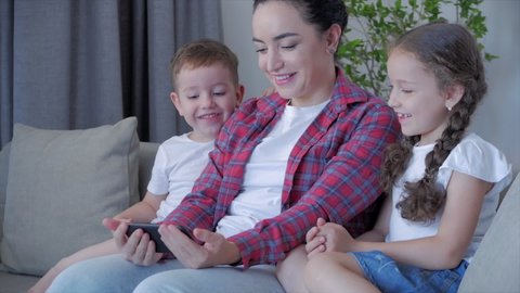 Happy family,mom and cute children playing with at home relaxing use a smartphone cuddling sit on sofa daughter and son laugh, watch funny videos.