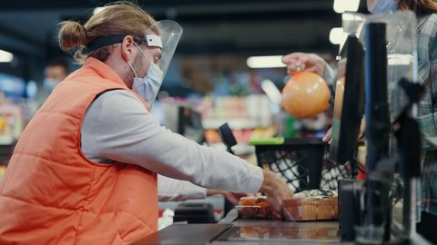 Bearded young cashier wearing respiratory mask face shield servicing young woman customer in supermarket grocery store during coronavirus quarantine.