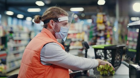 Portrait of young caucasian man cashier in face shield respiratory mask looking at camera sitting at cash resgiter servicing customers of supermarket.