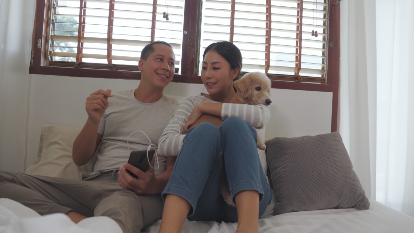 An Asian young couple listening to music on one headphone in the bedroom. They hug their dogs and are happy to listen to romantic music in the home