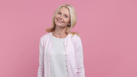 Smiling charming beautiful elderly gray-haired blonde woman lady 40s 50s years old wearing white blank casual t-shirt posing looking camera isolated on pastel pink color background studio portrait