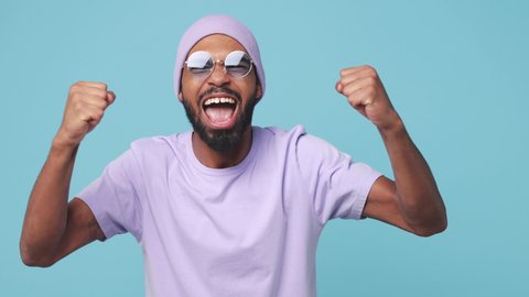 Worried excited happy young african american man 20s in violet t-shirt hat glasses isolated on blue turquoise background studio. People lifestyle concept. Doing winner gesture clenching fists say yes
