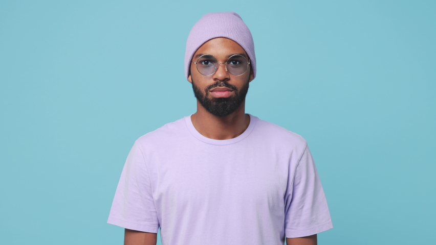 Smiling cheerful young bearded african american man 20s wearing violet t-shirt hat glasses posing isolated on blue turquoise color background in studio. People lifestyle concept. Looking at camera Royalty-Free Stock Footage #1062714160
