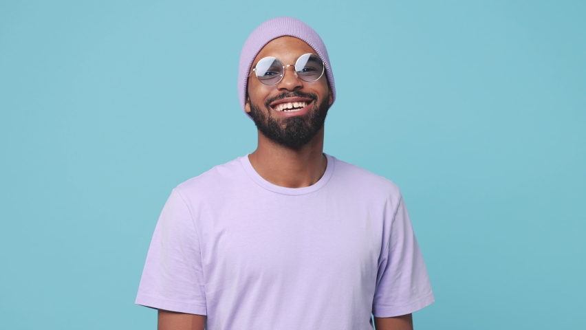 Smiling cheerful young bearded african american man 20s wearing violet t-shirt hat glasses posing isolated on blue turquoise color background in studio. People lifestyle concept. Looking at camera