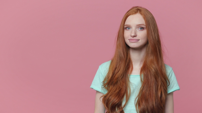 Smiling cheerful ginger redhead young woman wearing casual blue turquoise t-shirt posing isolated on pastel pink color background studio. People emotions lifestyle concept. Pointing index finger aside