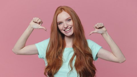 Cheerful excited redhead young woman in blue turquoise t-shirt posing isolated on pastel pink color background in studio. People lifestyle concept. Looking camera pointing thumbs on herself blinking