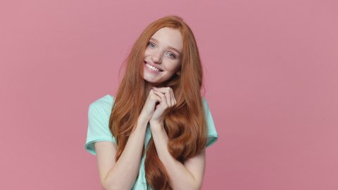 Smiling pretty ginger redhead young woman wearing casual blue turquoise t-shirt posing isolated on pastel pink color background in studio. People lifestyle concept. Looking camera put hands on heart