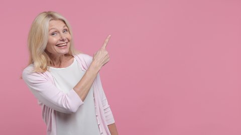 Smiling elderly gray-haired woman lady 40s 50s years old in white blank casual t-shirt pointing finger aside on workspace promo area mock up isolated on pastel pink color background studio portrait