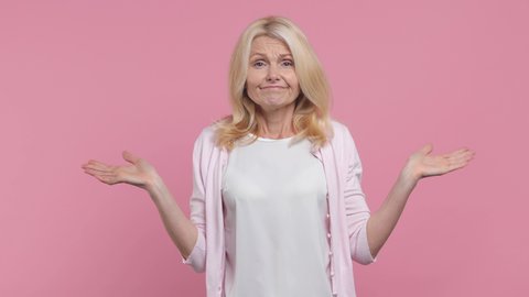 Confused perplexed elderly gray-haired blonde woman lady 40s 50s years old wearing white blank casual t-shirt looking camera spreading hands isolated on pastel pink color background studio portrait