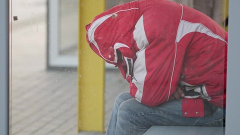 An unrecognizable man in a red jacket sits at a public transport stop. No face visible. A man sleeps on a bench at a bus stop. The tramp is resting in the street. A homeless man sleeps. Drunkard