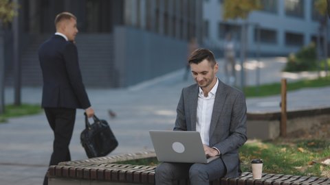 Young businessman in the city sits on bench, takes a laptop and works. Businessman typing on laptop computer outdoor. Man in suit working with laptop while sitting on bench. Remote work concept.