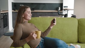 Smiling woman enjoying time watching video on smartphone and drinking tea or coffee lying on cozy sofa in living room. Young female resting on couch with hot drink and using mobile phone at home.
