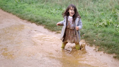 Happy little girl joyfully jumping and splashing in muddy puddles before running away. Young child having fun and getting dirty in puddles. Kids playing outside