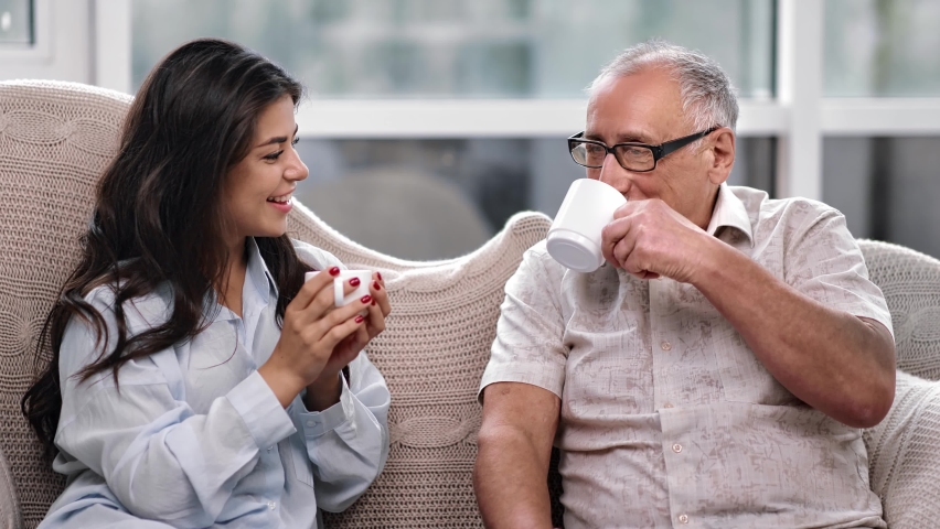 Smiling 70s grandfather father and young adult granddaughter daughter drinking tea and talking at home. Happy family relatives having friendship friendly conversation. Shot with RED camera in 4K Royalty-Free Stock Footage #1062728209