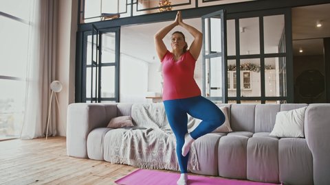 Young attractive plus size woman practicing yoga in static tree pose at home, hands in Namaste, full length