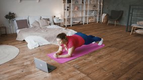 Sport training tutorial. Young overweight lady doing plank exercise, watching online fitness lessons on laptop at home