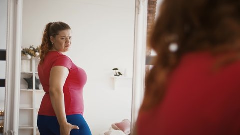 Body weight problem. Depressed lady with excess weight looking at mirror, feeling stressed about her figure and obesity
