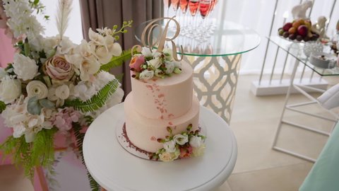 Delicious wedding cake with two floors decorated with pink details, roses and a topper in the shape of a Latin letter stands on background of buffet, slide of glasses of champagne and decor of flowers