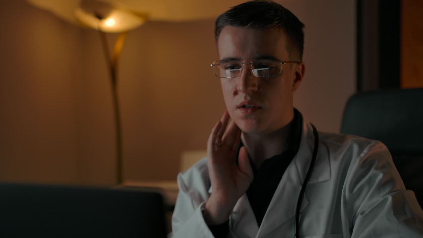 Male doctor shows how to probe lymph nodes through online communication. On-line conference and a conversation with patient from home. Clever Caucasian doctor in glasses and robe in cozy interior Royalty-Free Stock Footage #1062729307