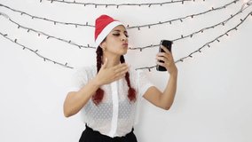 Young girl with a red christmas hat making a video call with a cell phone very happy, dancing, waving and sending kisses