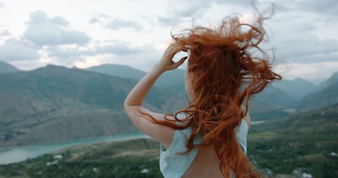 Gorgeous caucasian girl with red hair looking at scenic view from top of a mountain. Wind cinematically blowing hair and white dress - tranquility, peace, freedom 4k footage