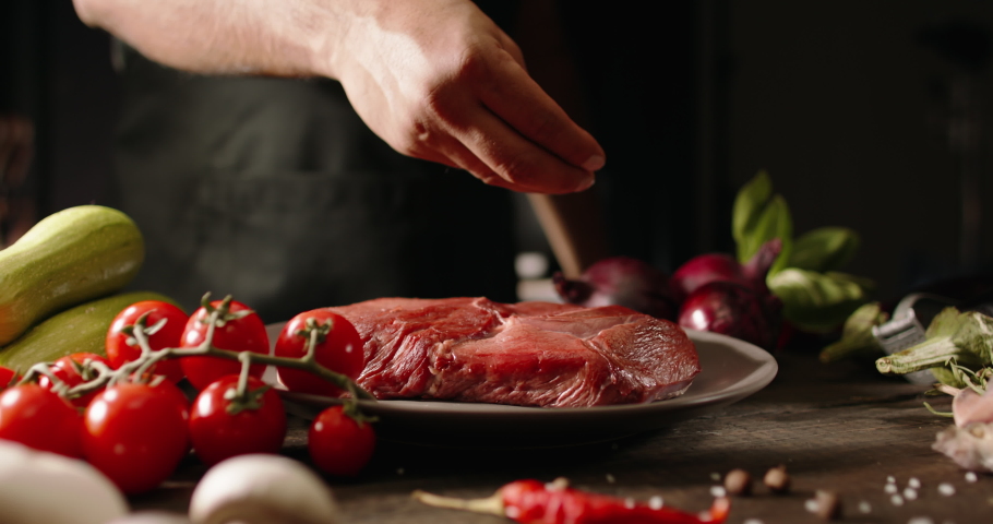 Chef applying grained salt on raw piece of steak. Cooker preparing meat on professional kitchen table with various vegetables 4k footage Royalty-Free Stock Footage #1062736174